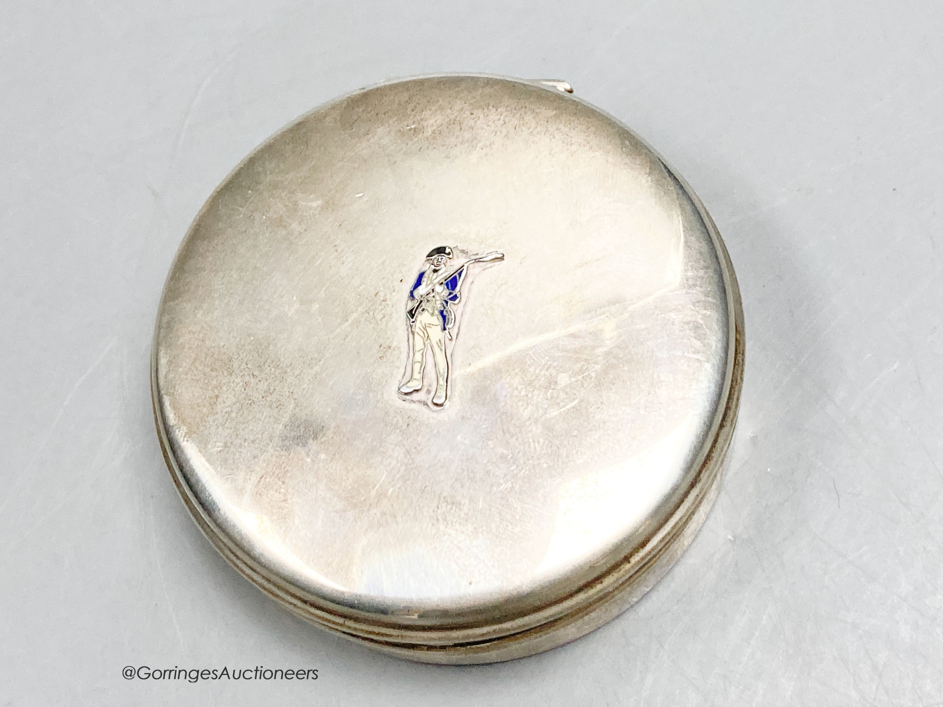 A white metal circular box and hinged cover, with enamelled applique modelled as a soldier, stamped sterling and bearing the stamp 'Cartier', diameter 73mm, gross weight 98 grams.